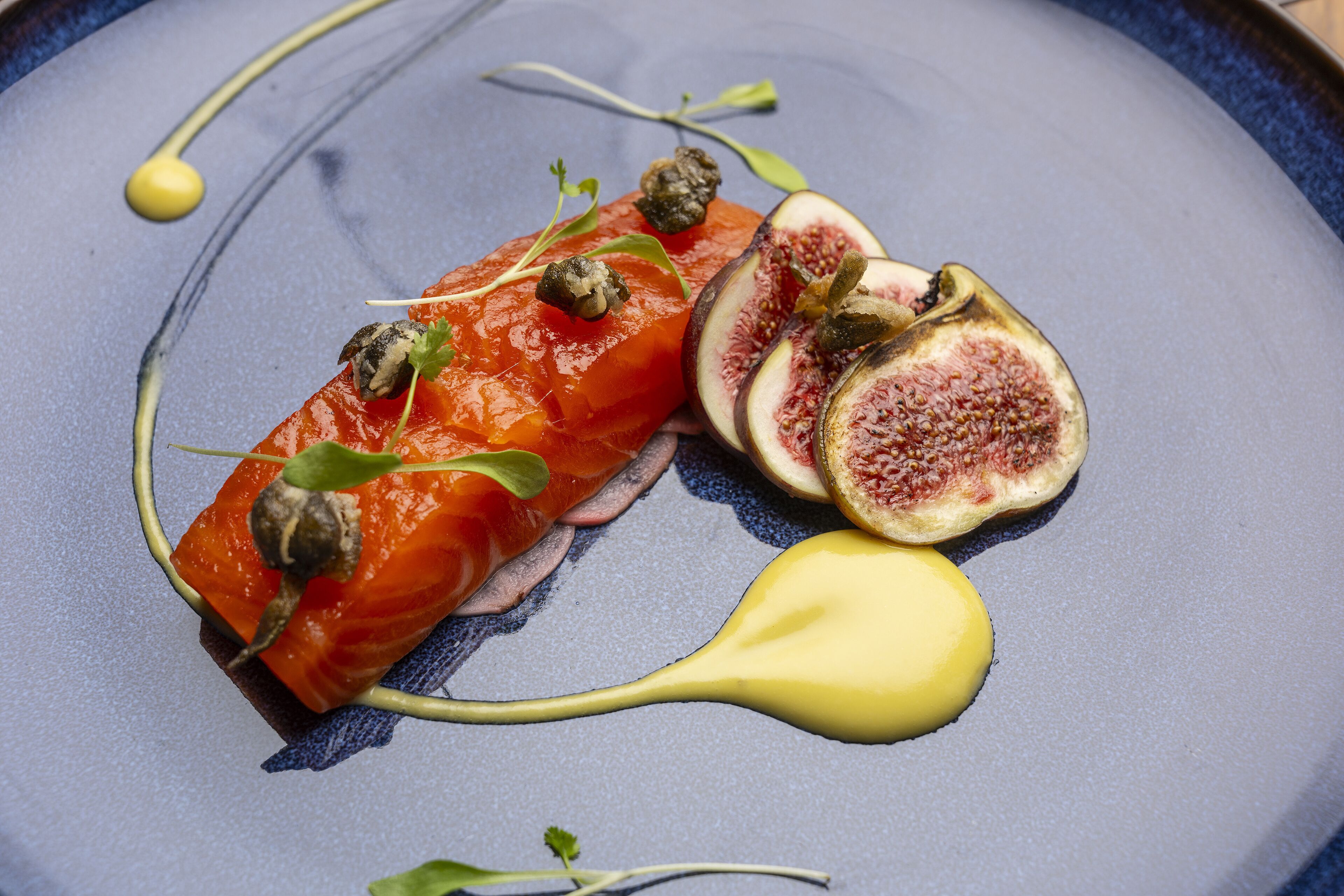 A dish with salmon, figs, and a yellow sauce on a blue plate.