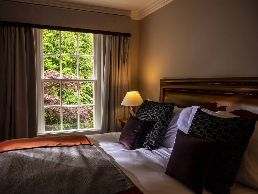 Camera d'albergo con una grande finestra, un letto con molti cuscini, una lampada e vista sul verde.