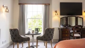 A hotel room with a bed, two upholstered chairs, a small table with a kettle and cups, a desk with a mirror, and a TV mounted on the wall.