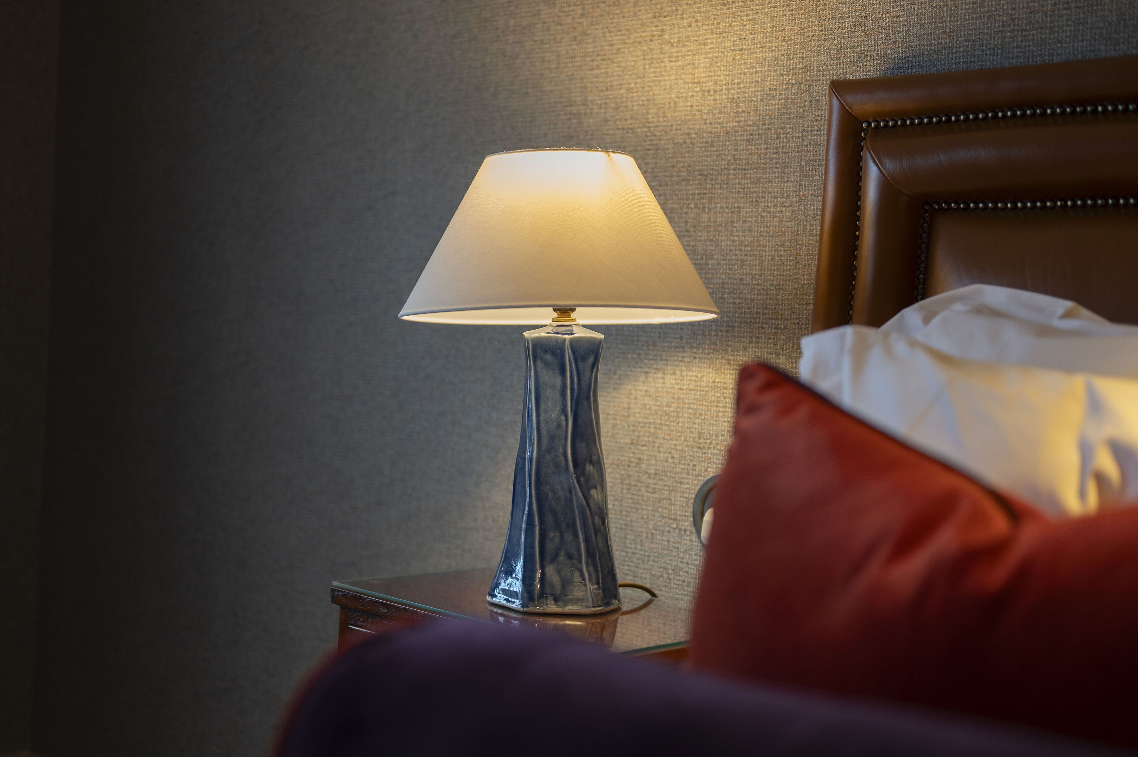 A bedside table with a lamp, a bed with a headboard, and a red pillow.