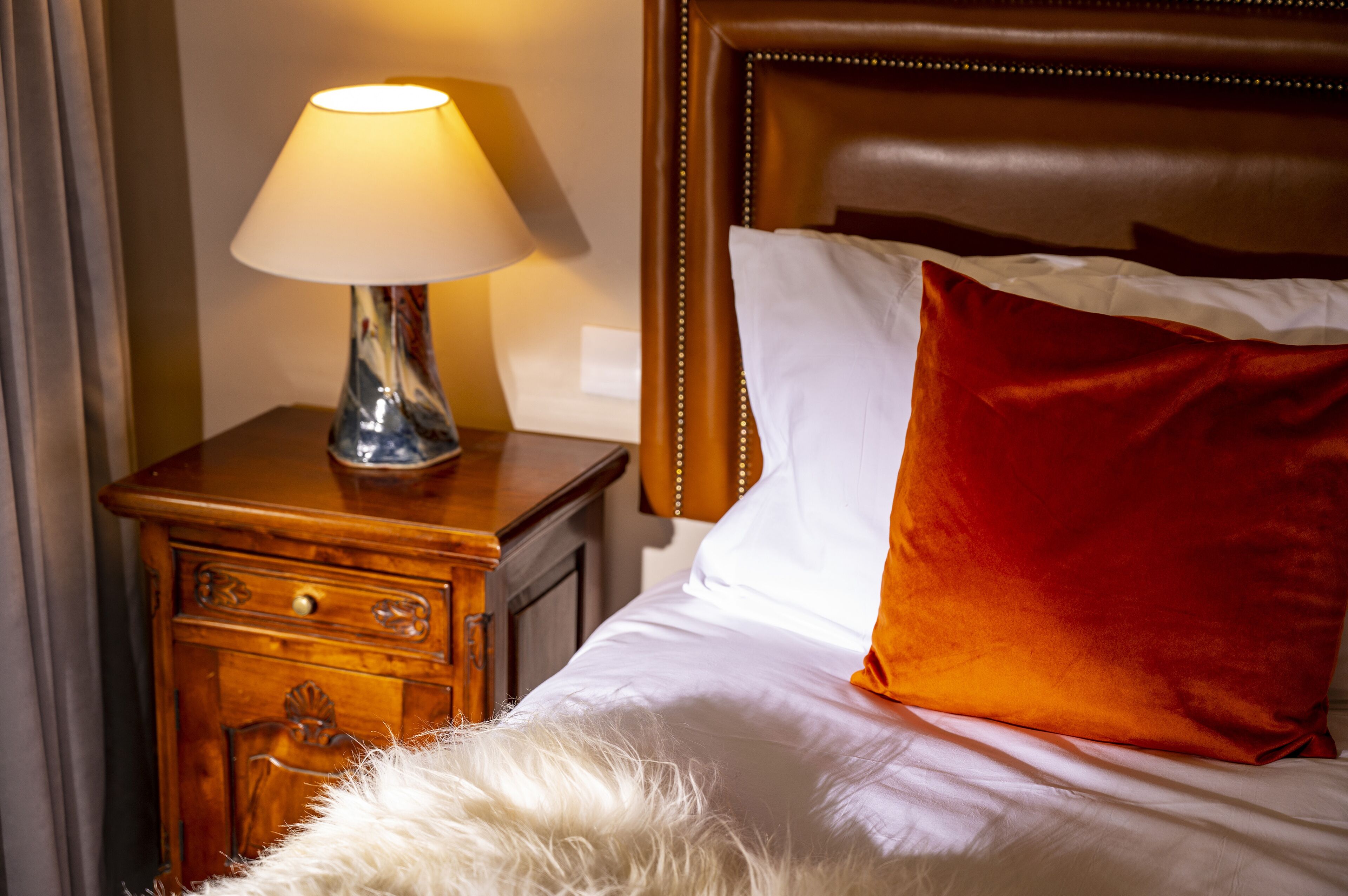 Een net opgemaakt bed met witte en rode kussens, een zacht, wit kussen en een lamp op een houten nachtkastje.