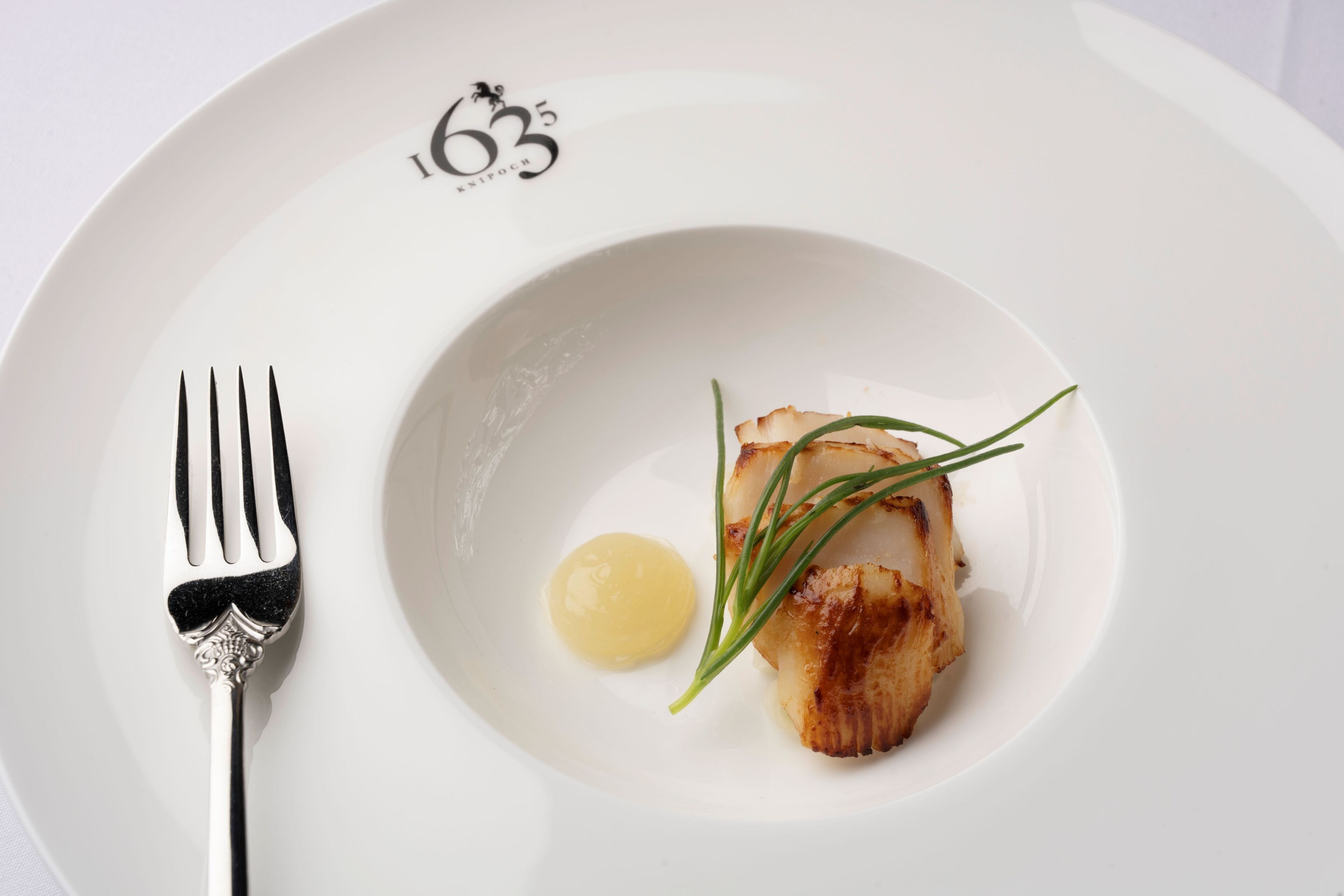 Un piatto bianco con un filetto di pesce scottato, una quenelle di burro e un guarnizione di erba cipollina.