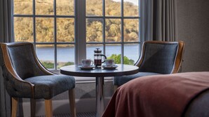 A room with two chairs facing a window overlooking a lake, a small table with two cups and a pitcher, and a bed with a red blanket. - Knipoch House Hotel (Oban)