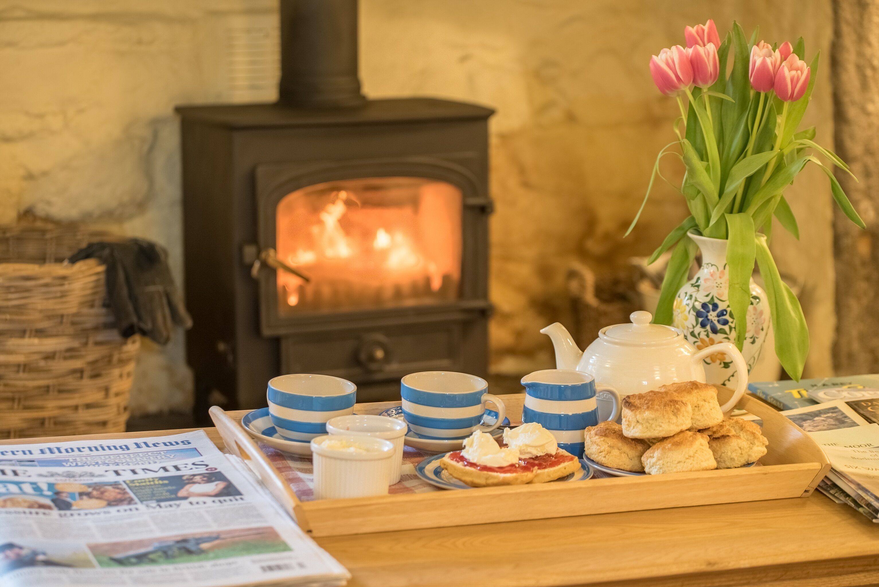 Une pièce chaleureuse avec un poêle à bois, une table avec un journal, un vase rempli de fleurs et un plateau avec des biscuits et de la crème.