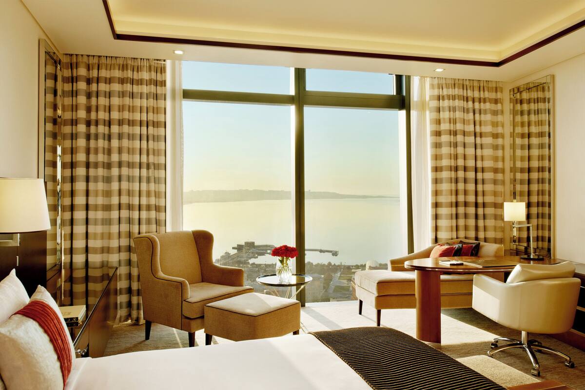 Ein Hotelzimmer mit großem Fenster, einem Bett, zwei Sesseln, einem kleinen Tisch und einem Schreibtisch mit Stuhl.
