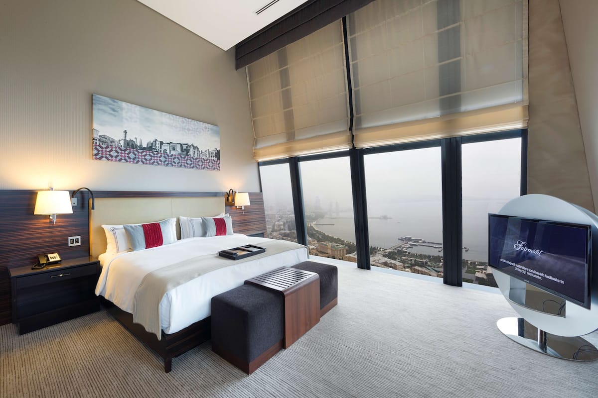 Ein modernes Hotelzimmer mit einem großen Bett, einem Fernseher, einem Nachttisch und Blick auf eine Stadtkulisse durch große Fenster.