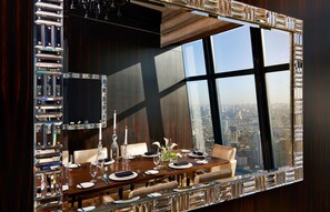 A high-rise dining area with a panoramic city view, a large mirror, and a modern chandelier.