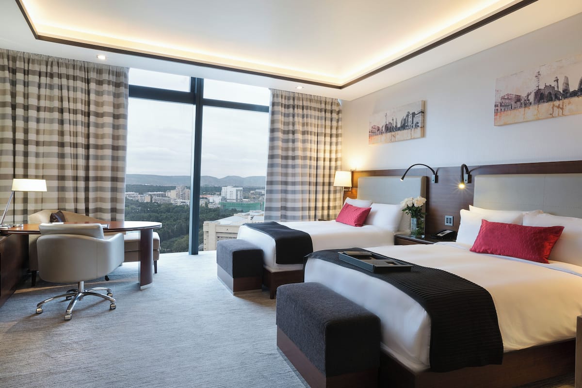 Ein Hotelzimmer mit zwei Betten, einem Schreibtisch, einem Stuhl und großen Fenstern mit Blick auf die Stadt.