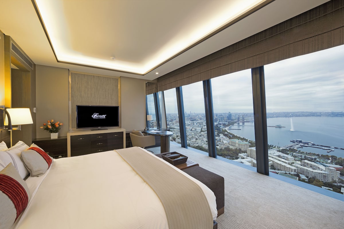 Ein modernes Hotelzimmer mit einem großen Bett, einem Fernseher, einem Schreibtisch und Blick auf Stadt und Wasser.