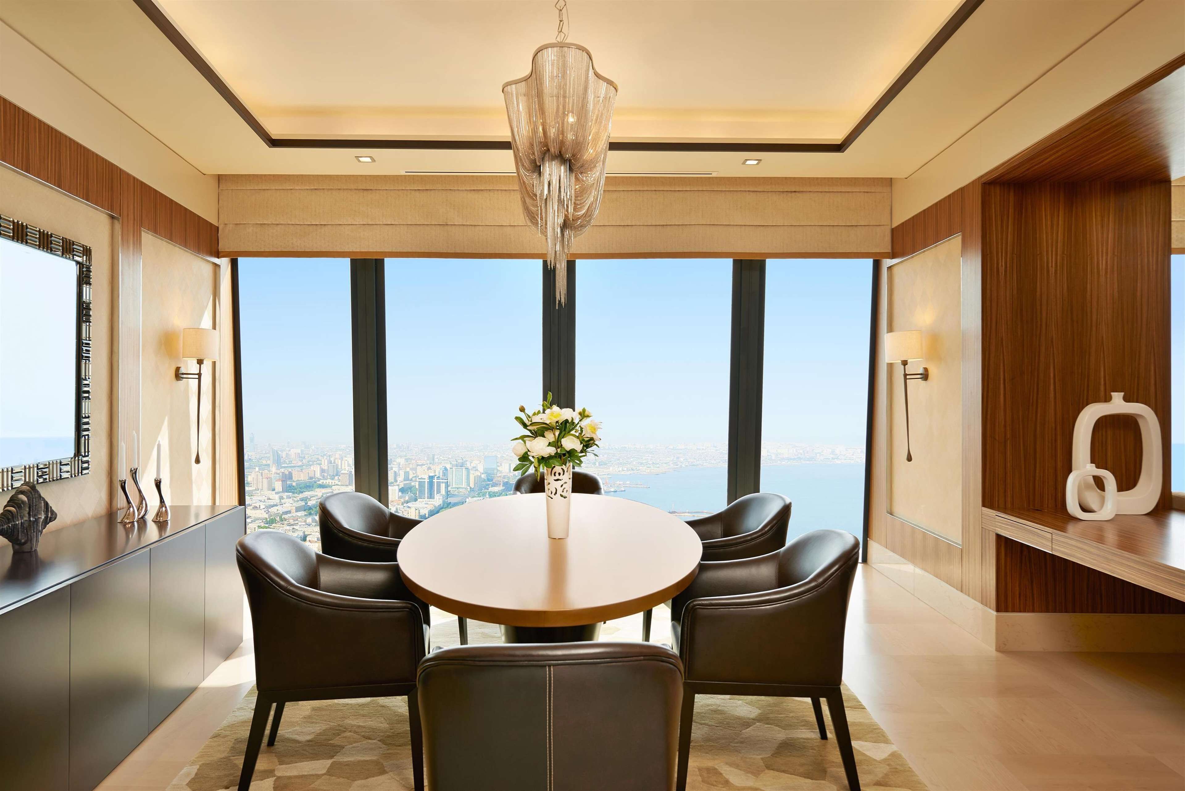 A modern dining area with a round table, four chairs, a chandelier, and a view of the city through large windows.