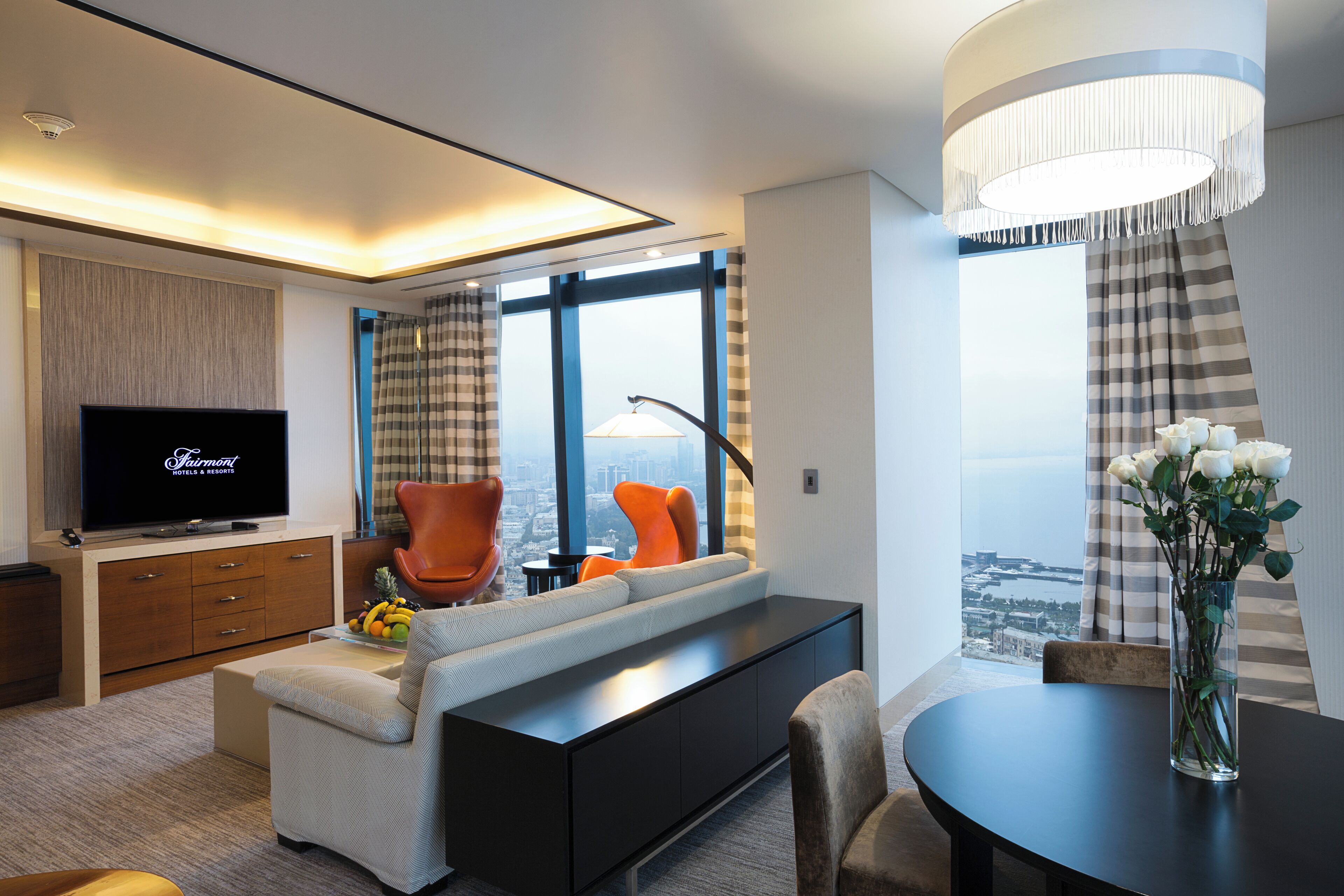 Ein modernes Hotelzimmer mit einem großen Flachbildfernseher, einer Couch, einem Esstisch und Blick auf die Stadt durch große Fenster.