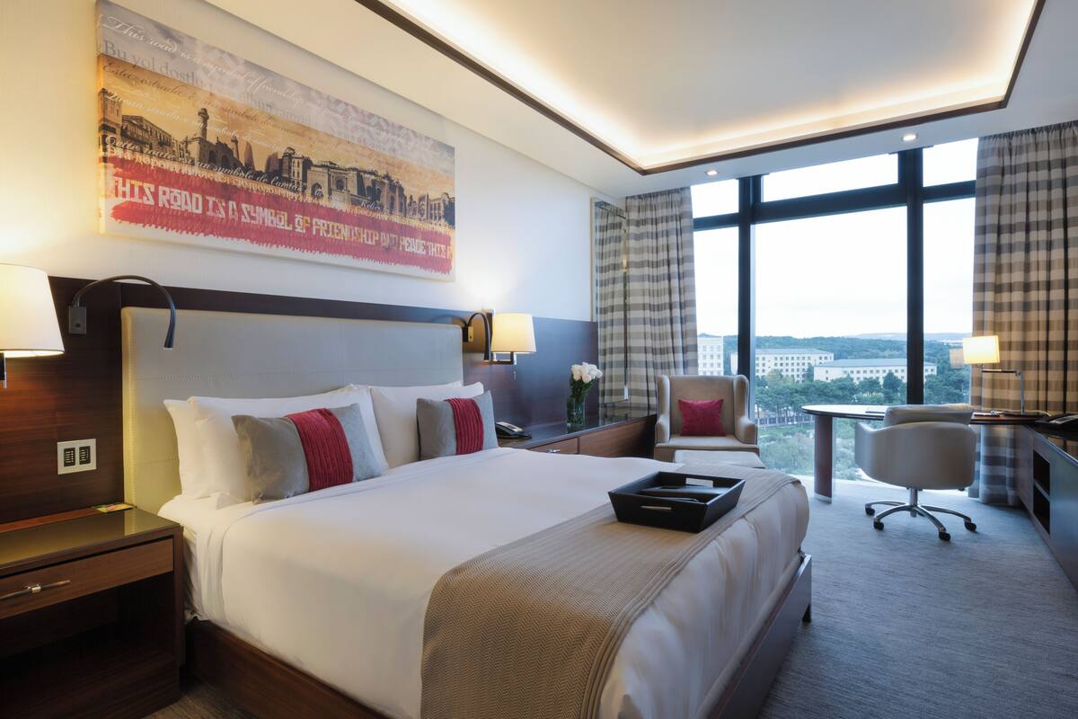 Ein Hotelzimmer mit einem großen Bett, einem Schreibtisch, einem Stuhl und Blick auf die Stadt durch große Fenster.