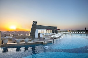 Una piscina infinita en la azotea con vista a la ciudad al atardecer, sillones de descanso y un pequeño edificio con techo inclinado.