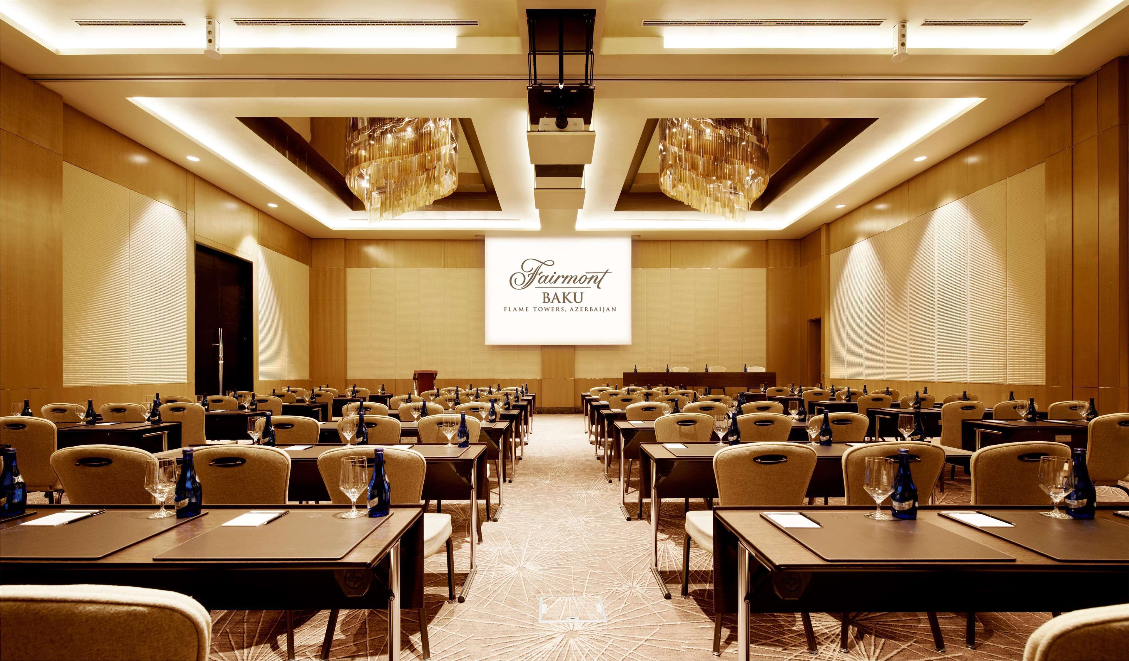 Een vergaderzaal met rijen tafels en stoelen, een groot scherm waarop 'Fairmont Baku Flame Towers, Azerbaijan' staat, en een moderne kroonluchter.