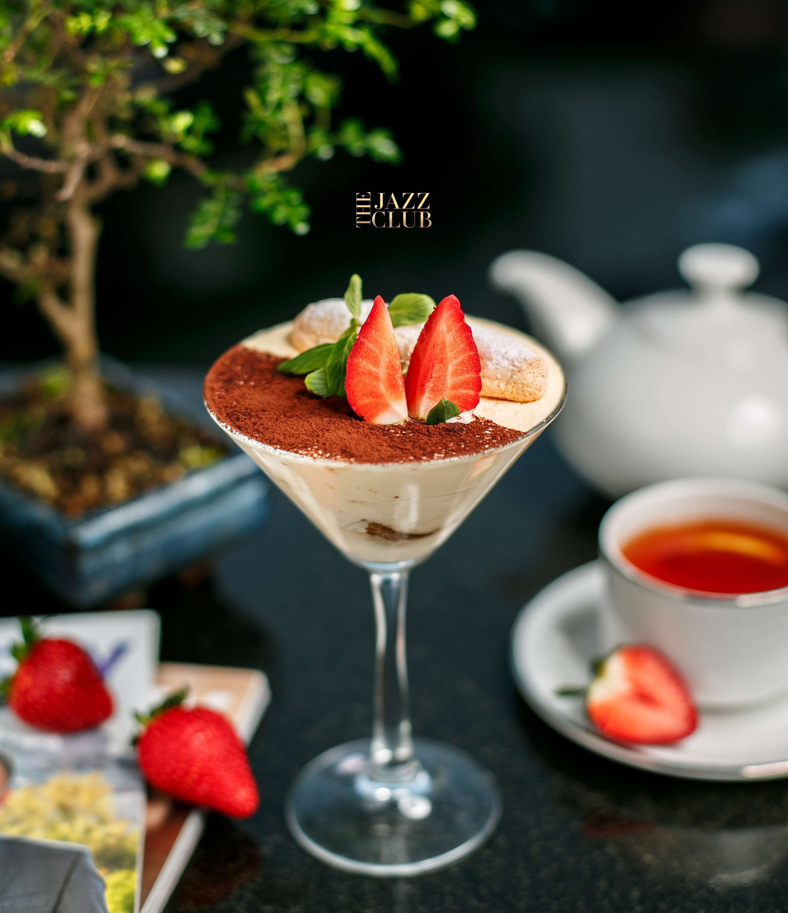 Een dessert in een martini-glas met aardbeien en cacaopoeder, een theepot, en aardbeien op een tafel.