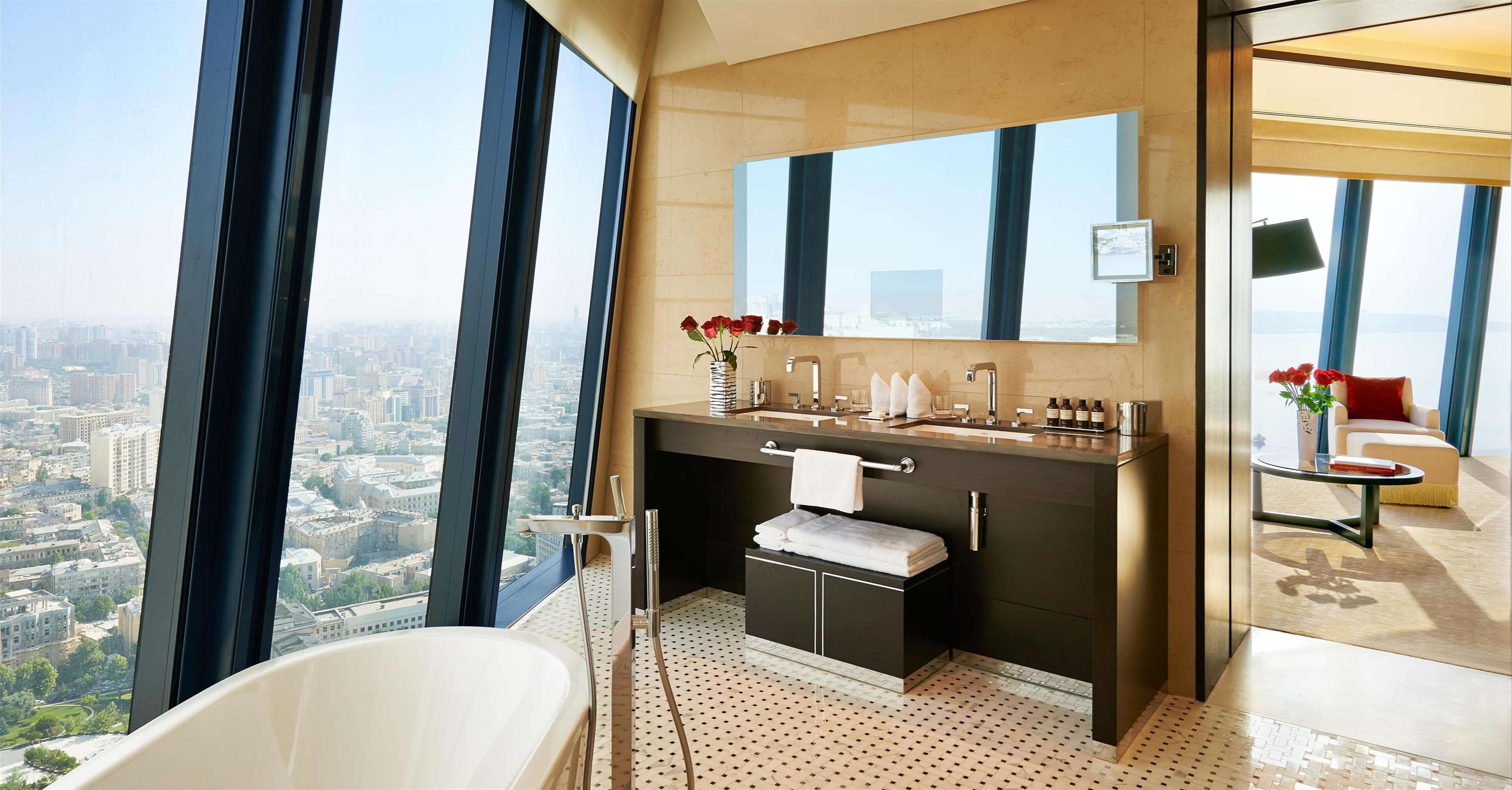 Un bany modern amb una banyera gran, un lavabo amb mirall i vista a un paisatge urbà a través de grans finestres.