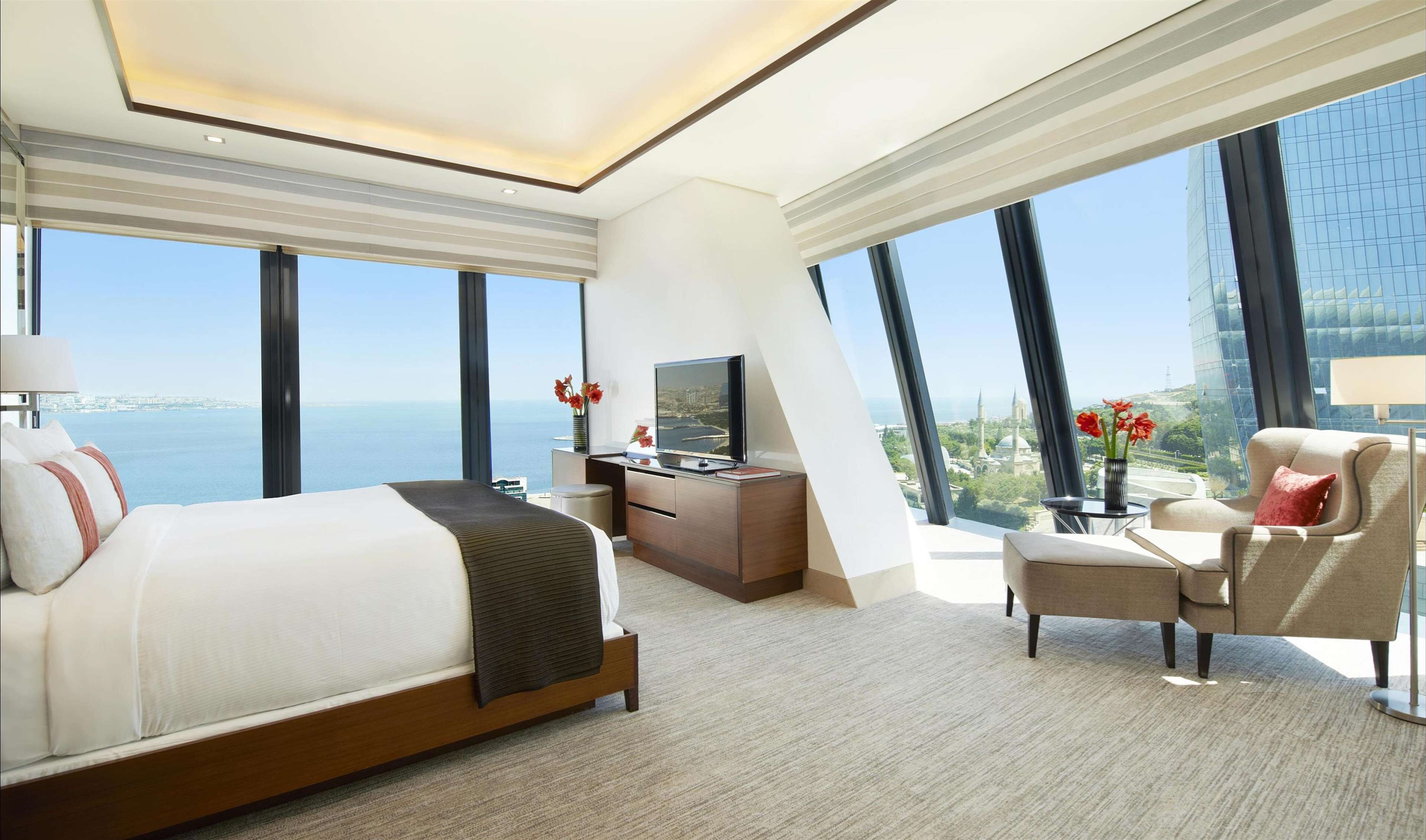 A modern hotel room with a large bed, a desk, a chair, and a TV. The room has large windows offering a view of the city and the sea.