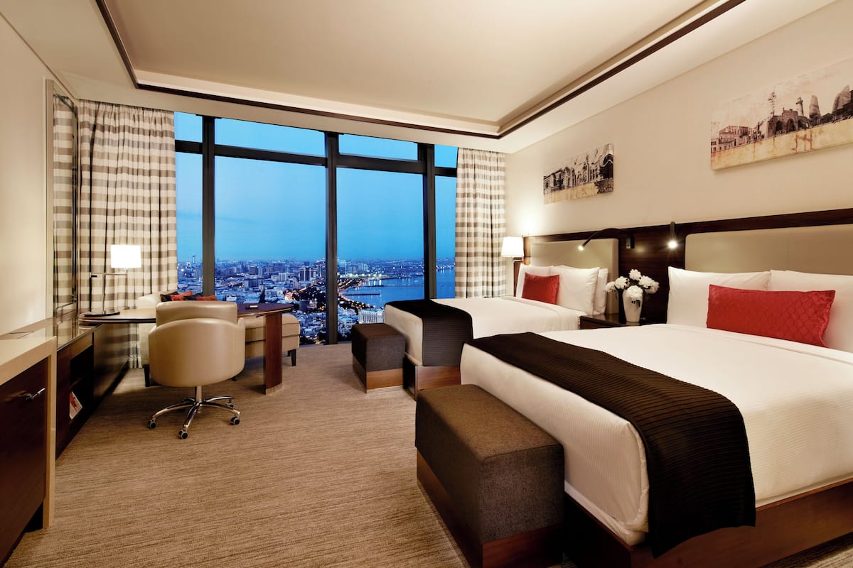 Ein Hotelzimmer mit einem großen Bett, einem Schreibtisch, einem Stuhl und Blick auf eine Stadtlandschaft durch große Fenster.