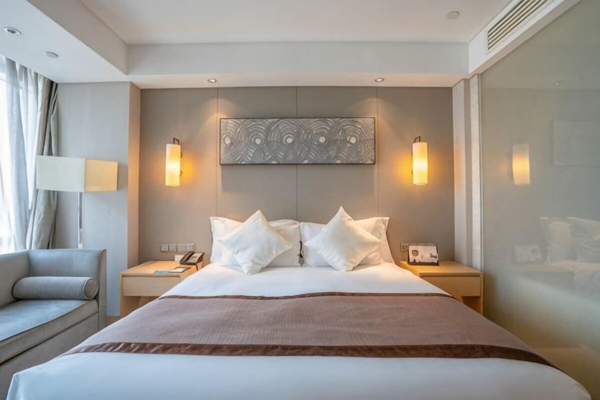 A hotel room with a large bed, two bedside tables, a nightstand, a lamp, and a view of the outside.