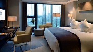 Une chambre d’hôtel moderne avec un grand lit, un bureau et une chaise. On y trouve deux lampes et une vue sur la ville grâce à de grandes fenêtres.