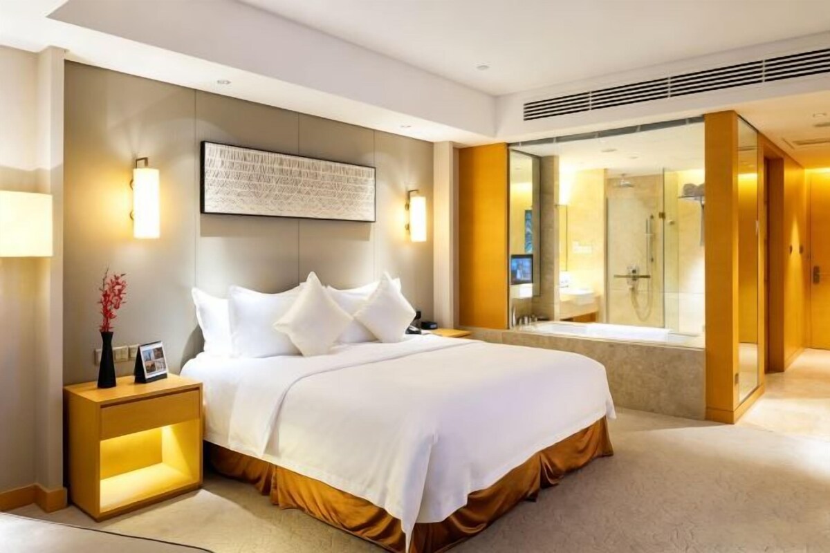 A modern hotel room with a large bed, bedside lamps, a nightstand, a framed artwork, and a bathroom with a glass shower enclosure.