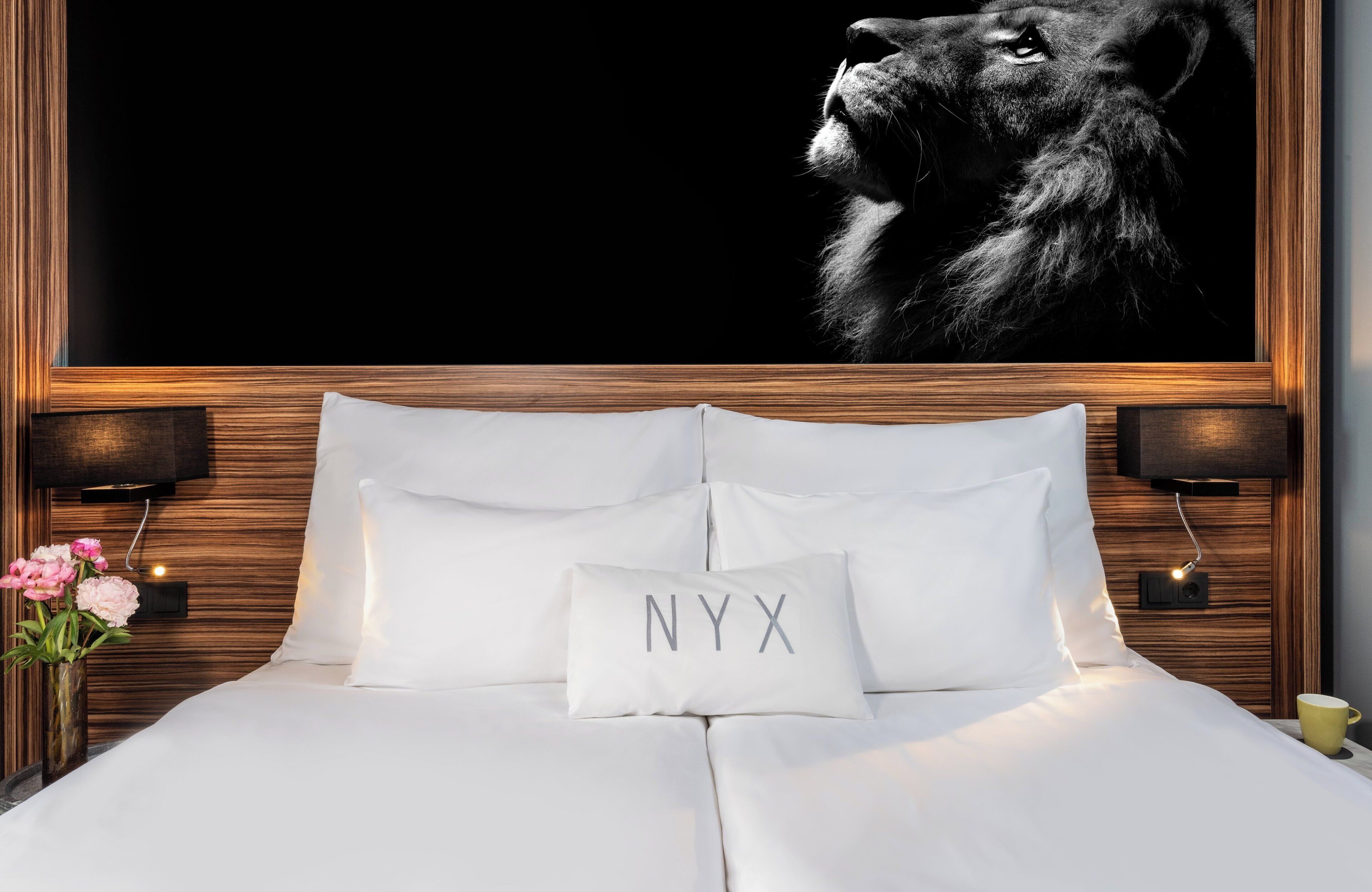A hotel room with a large framed picture of a lion, a bed with white bedding, a pillow with "NYX" on it, and two bedside lamps.