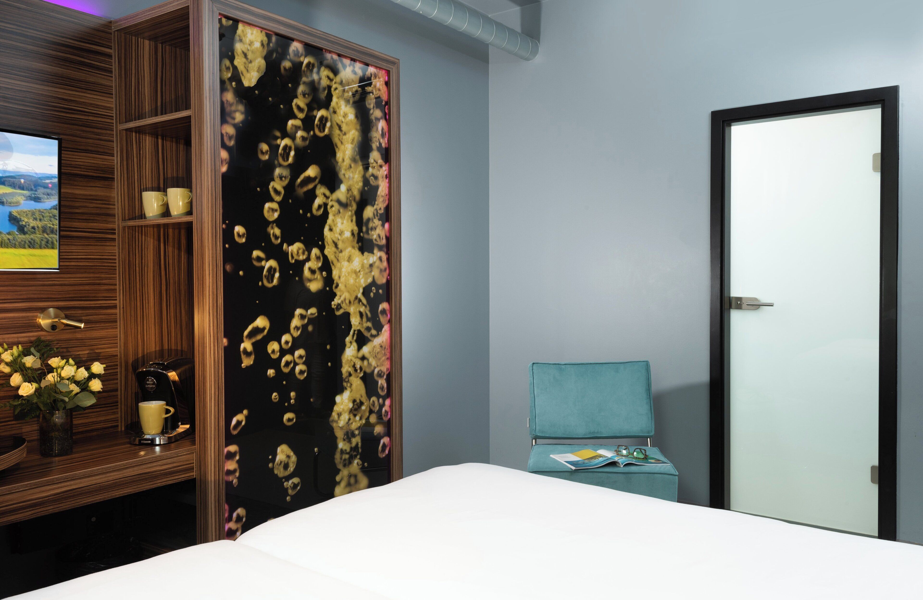 A hotel room with a bed, a wardrobe, a small table with a vase of flowers, a coffee machine, and a television displaying a scenic landscape.