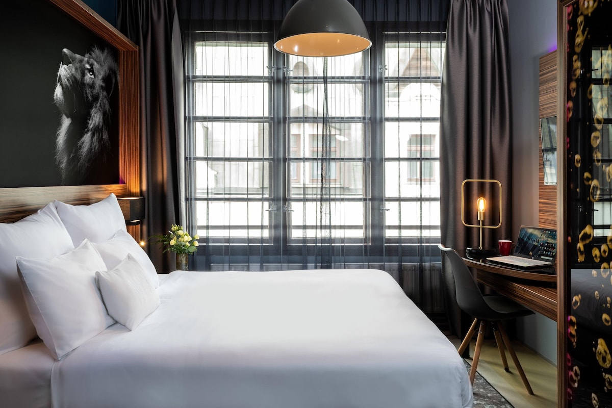 Habitació d'hotel amb un llit gran, una taula de treball amb un ordinador portàtil, una làmpada i una finestra amb cortines.