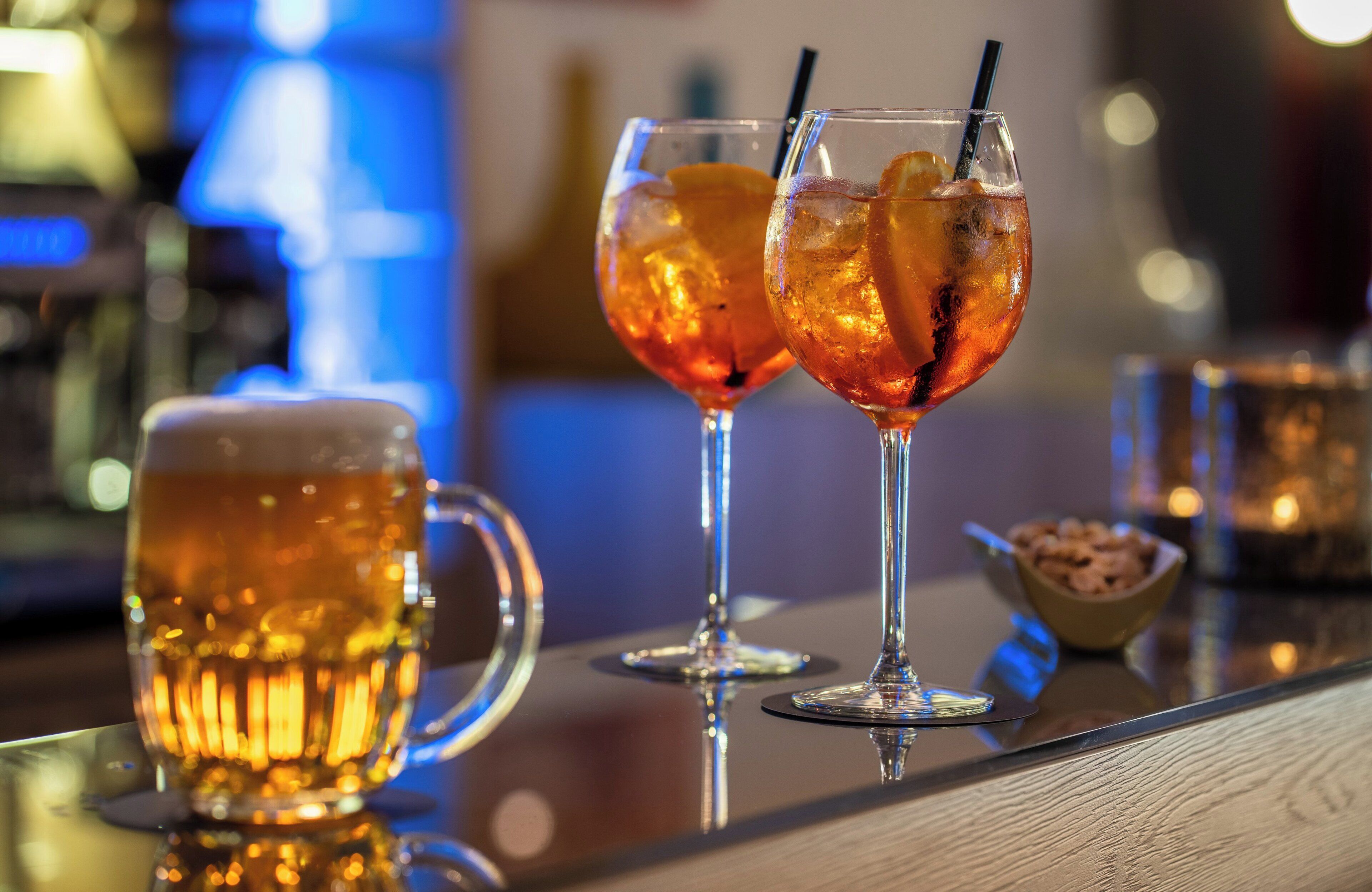 Dos vasos de bebida color ámbar con hielo y una pajita, y una jarra grande de cerveza sobre una superficie reflectante.