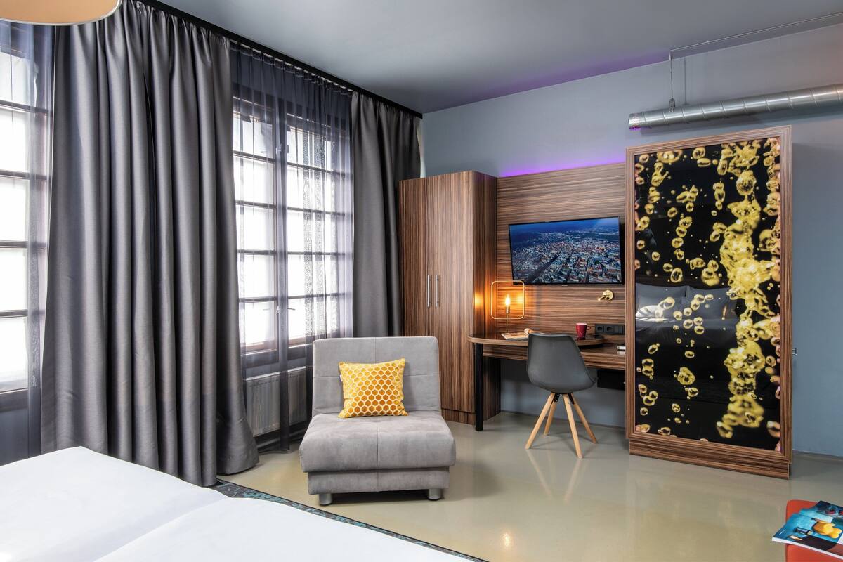 A modern hotel room with a large bed, a grey armchair with a yellow pillow, a desk with a computer, a TV, and a decorative wall panel.