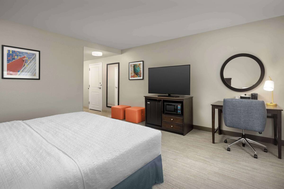 Habitación de hotel con cama, escritorio con silla, televisor, microondas y dos otomanas naranjas.