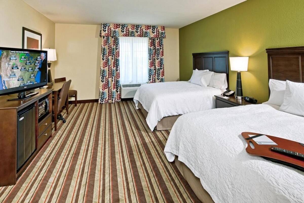 Habitación de hotel con dos camas, televisión, escritorio con lámpara y ventana con cortinas.