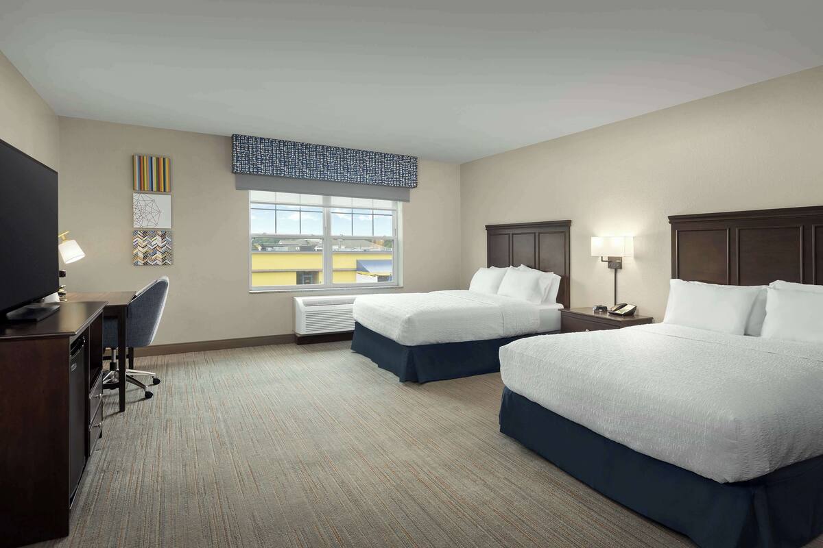 Habitación de hotel con dos camas, un escritorio con silla, televisor y ventana con vistas a edificios.