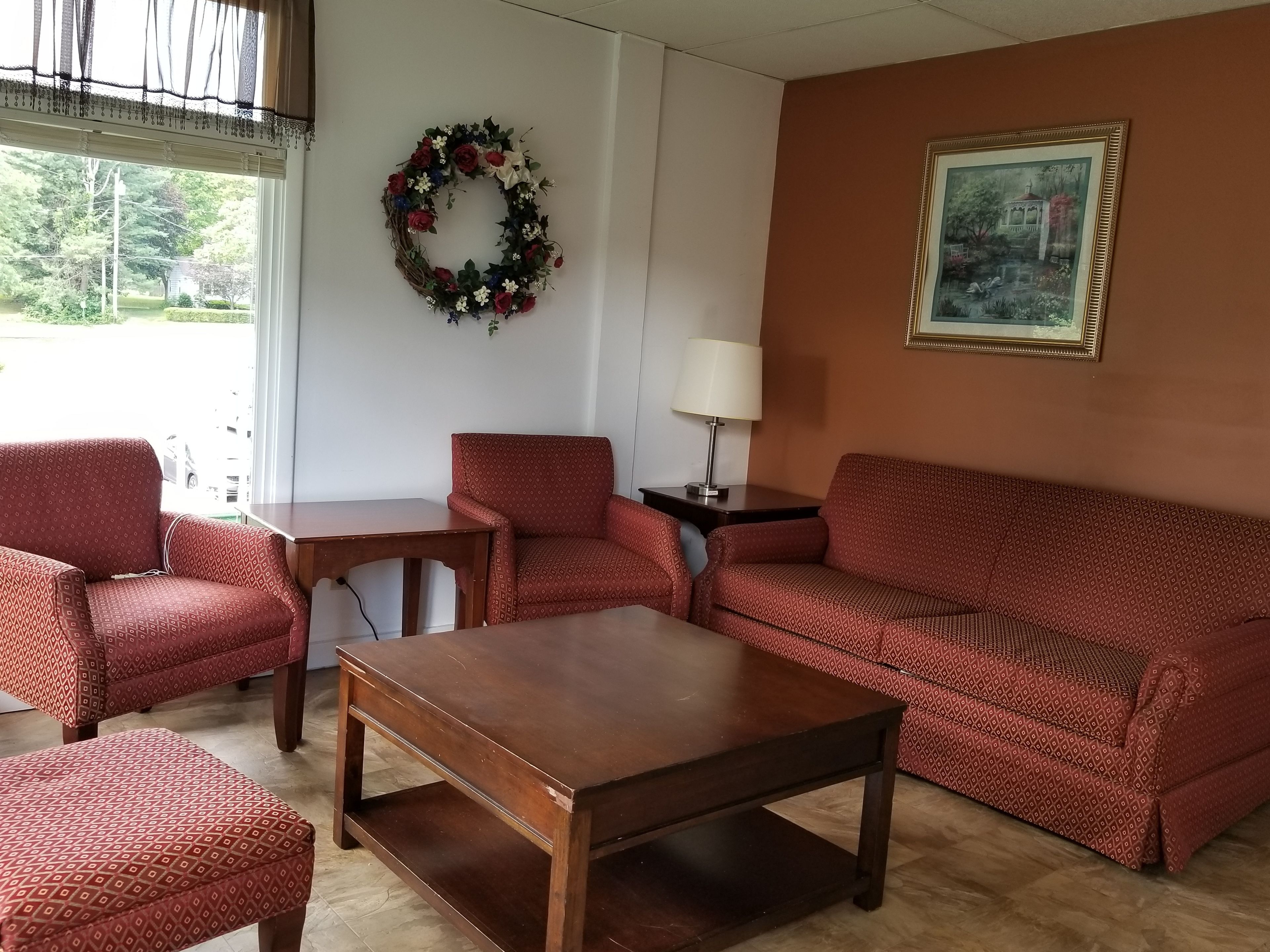 A living room with a wooden coffee table, two patterned armchairs, a sofa, a small table, a lamp, a framed painting, and a wreath on the wall.