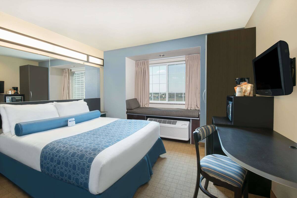 Habitación de hotel con cama, escritorio, silla, televisión, ventana con cortinas y espejo.