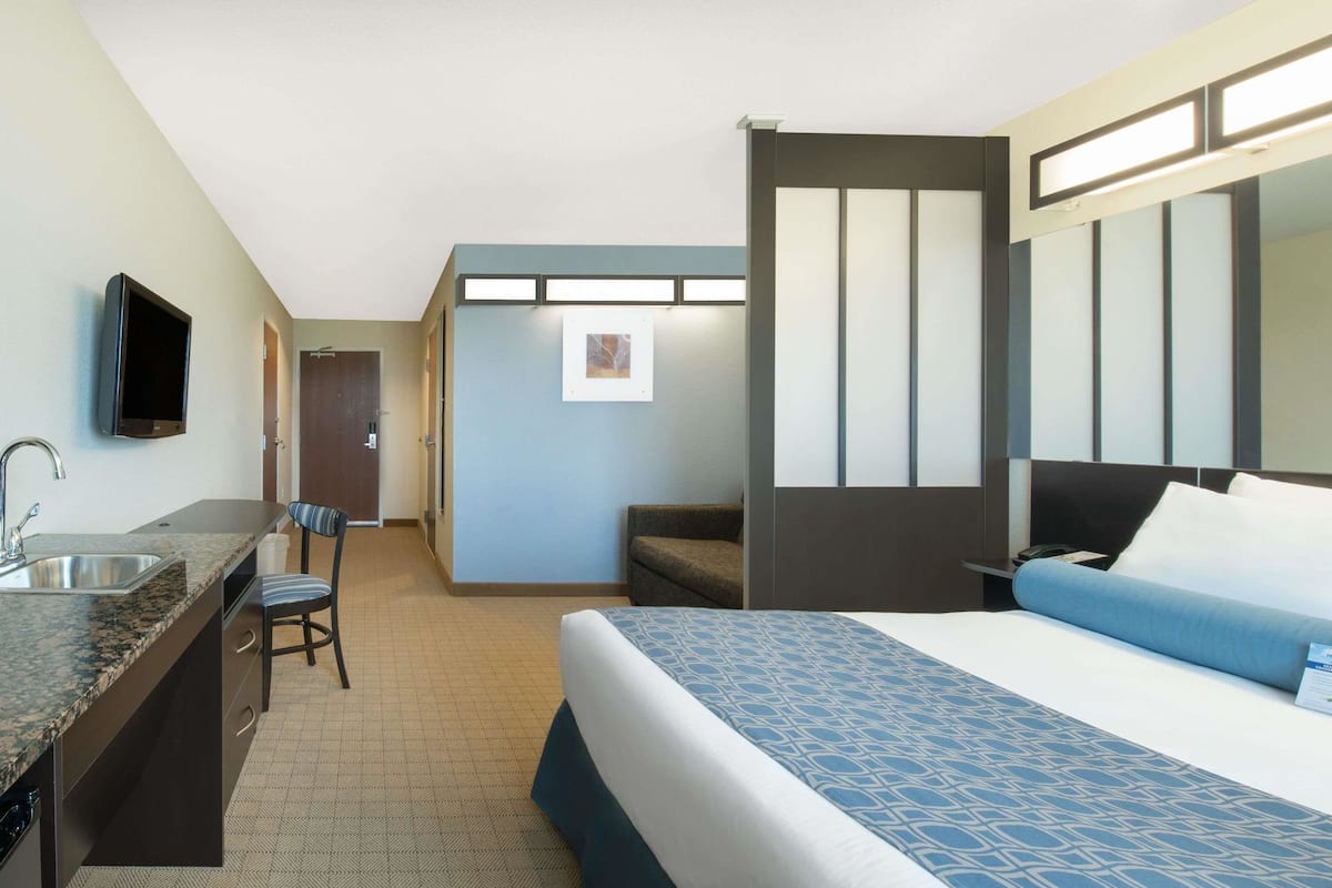 Habitación de hotel con una cama, un escritorio con lavamanos, una silla, un televisor y una puerta que da a otra habitación.