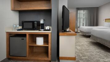 Habitación de hotel con kitchenette integrada, televisión de pantalla plana, cama con ropa blanca y ventana con cortinas.