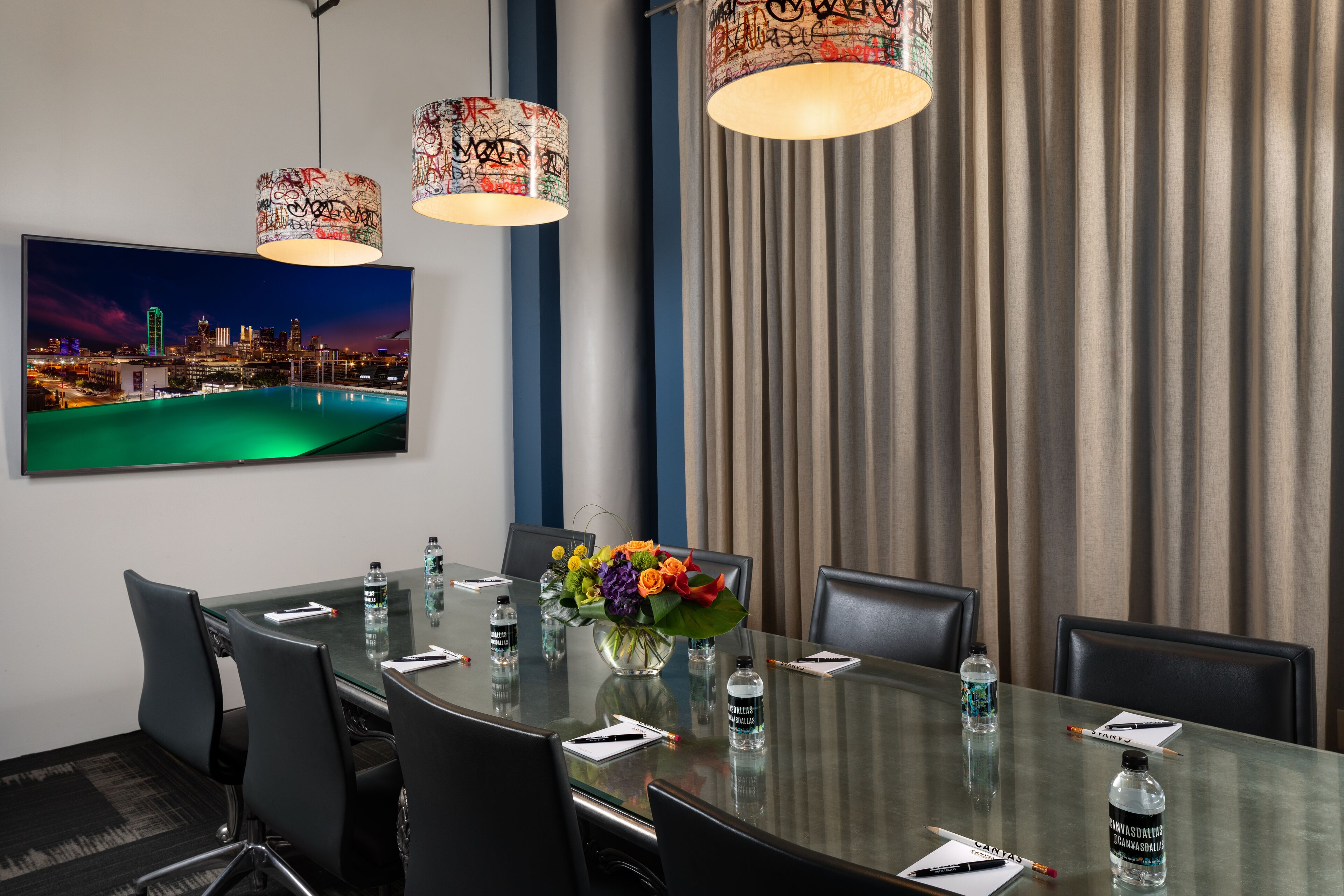 A conference room with a glass table, black chairs, a floral centerpiece, water bottles, and a cityscape painting on the wall.