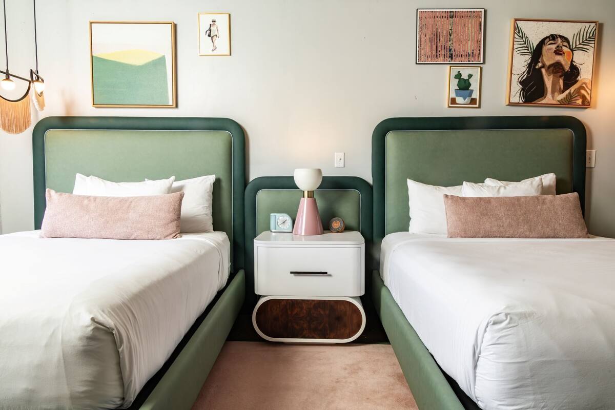 A room with two beds, a bedside table with a lamp, a clock, and framed artwork on the wall.