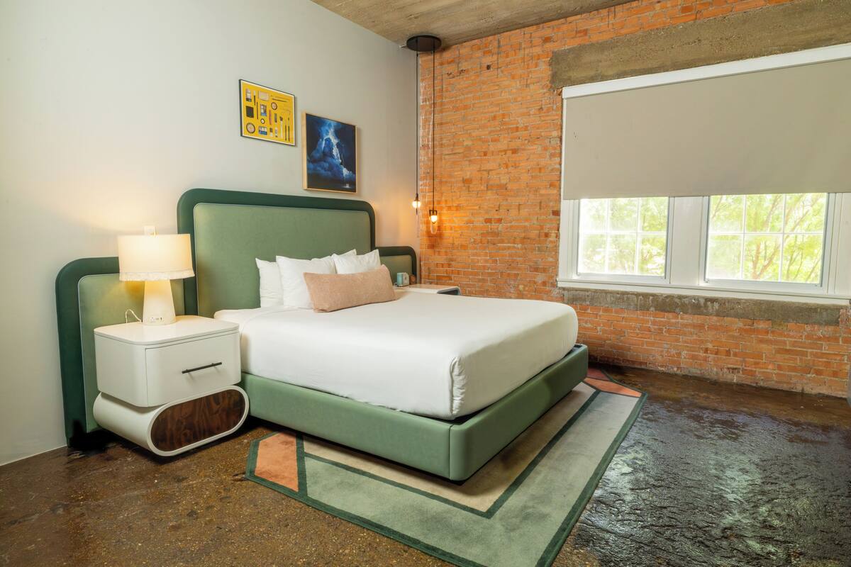 A bedroom with a green bed, white bedding, a nightstand with a lamp, a brick wall, and a window.