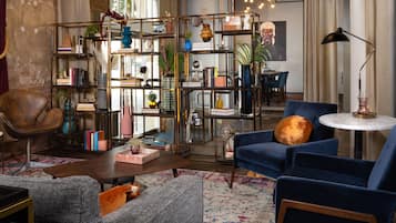 A cozy living room with a blue sofa, a grey armchair, and a wooden coffee table. There is a bookshelf filled with books and decorative items, and a large window providing natural light.
