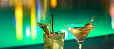 A cocktail glass with a lime garnish and a tall glass with a mint garnish, both on a table by a poolside.