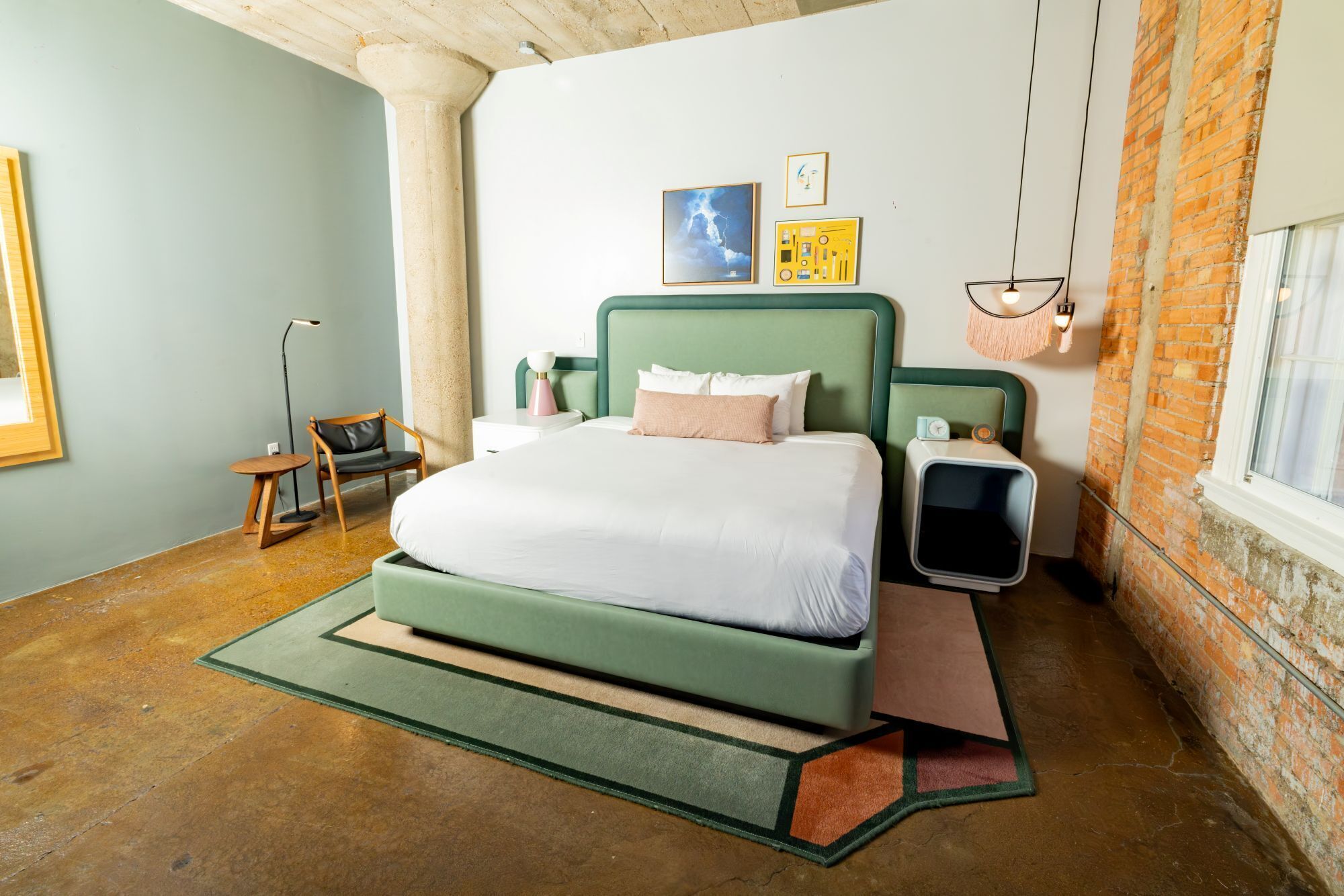 Un dormitorio moderno con una cama verde, una mesita de noche pequeña, una silla, una lámpara de pie y una pared de ladrillo.