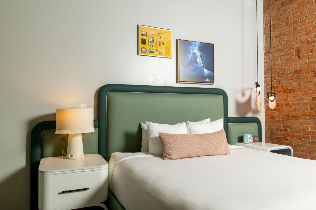 A hotel room with a green headboard, a bedside table with a lamp, a bed with white and pink pillows, and two framed pictures on the wall.