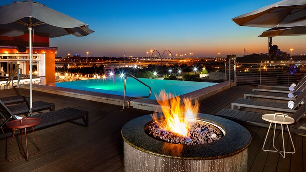 A rooftop pool area with lounge chairs, a fire pit, and a view of a city skyline at dusk. - Canvas Hotel Dallas (Dallas)