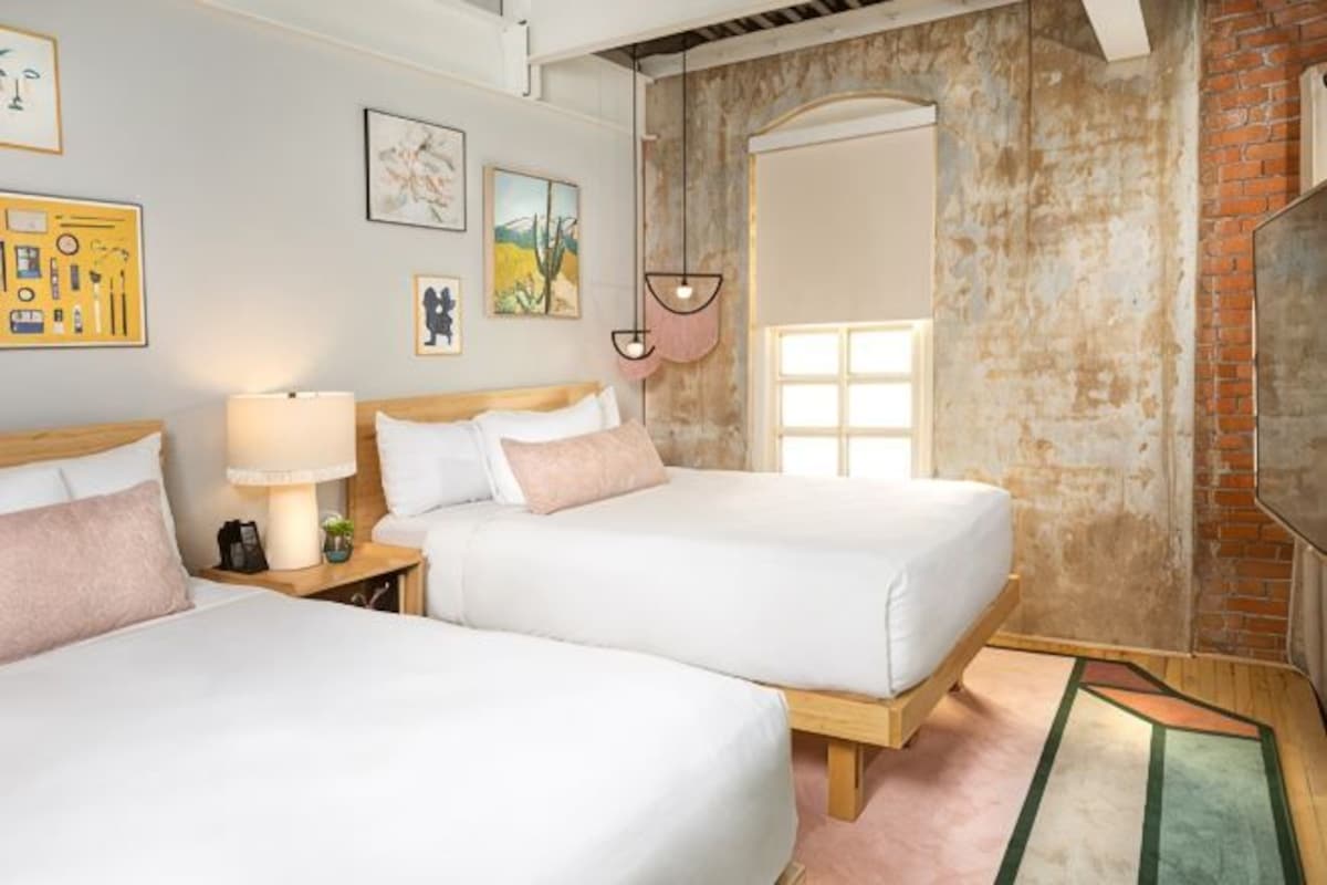 A room with two beds, a lamp, artwork on the walls, and a brick wall.