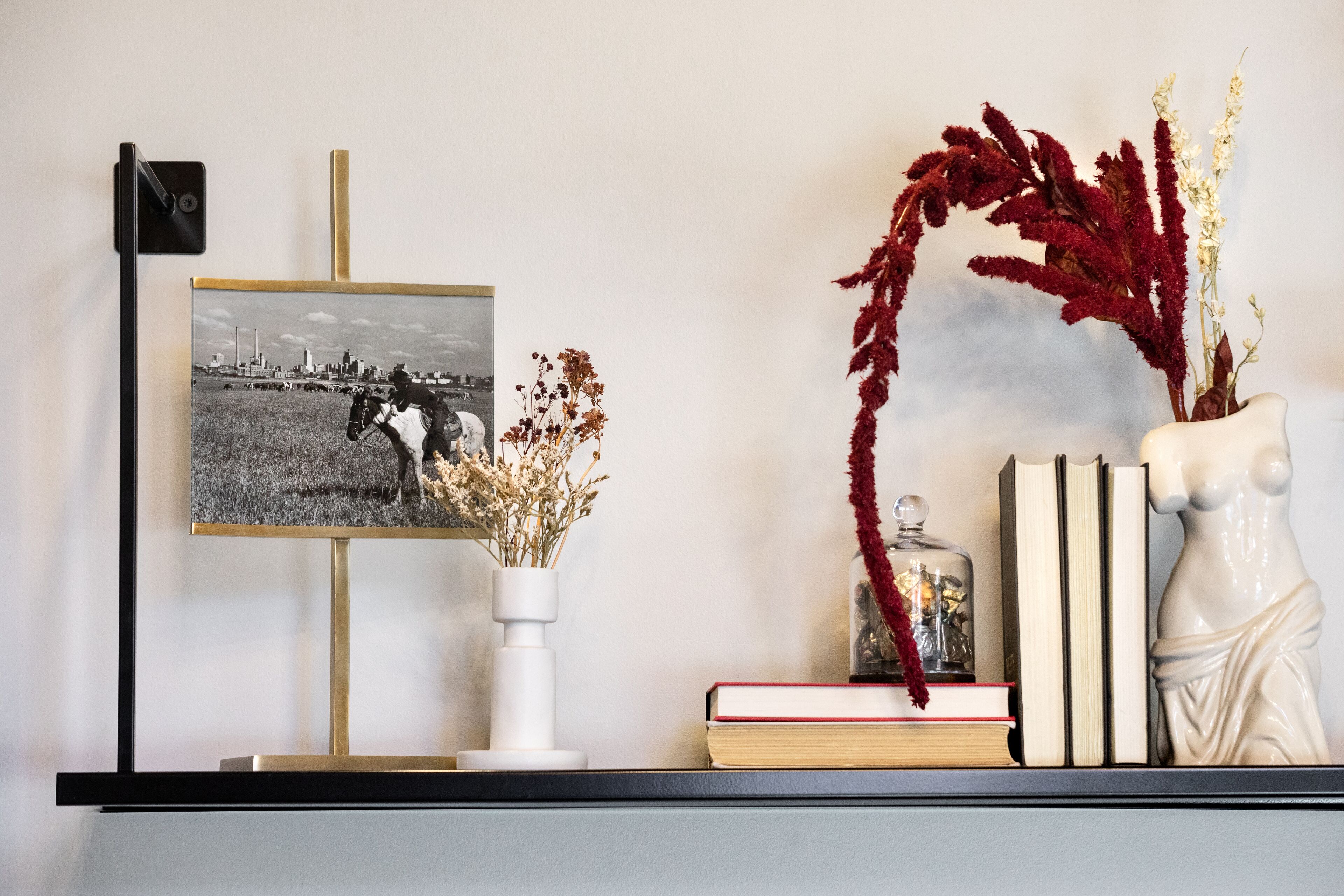 Una estantería con una fotografía blanco y negro enmarcada, un jarrón blanco con flores secas, un frasco de vidrio con un lazo rojo y una escultura de busto.