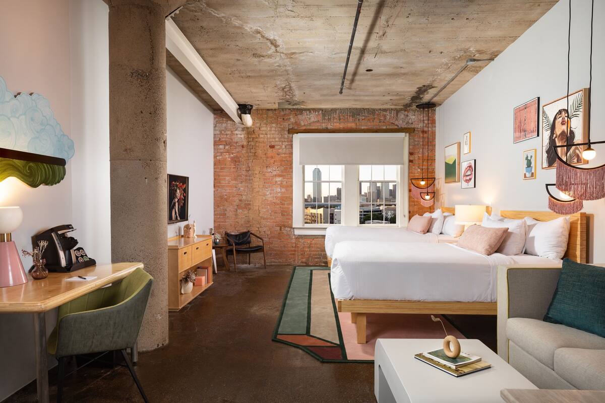 A modern hotel room with a brick wall, a large bed, a desk with a chair, and a view of the city through a window.
