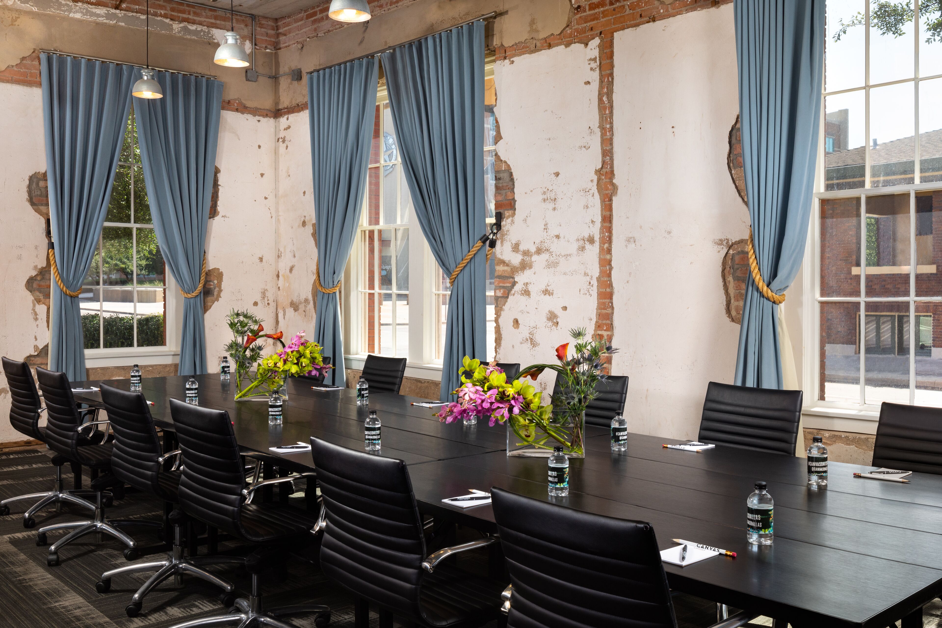 Una sala de conferencias con una larga mesa, sillas negras y cortinas azules. Hay botellas de agua y un jarrón con flores sobre la mesa.