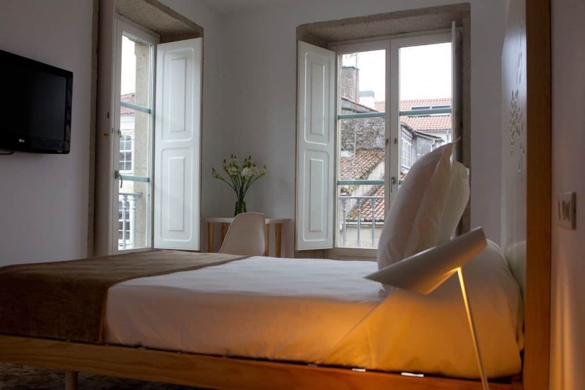 Ein Schlafzimmer mit einem Bett, einer Nachttischlampe, einem Fernseher und zwei offenen Fenstern mit Blick auf Gebäude.
